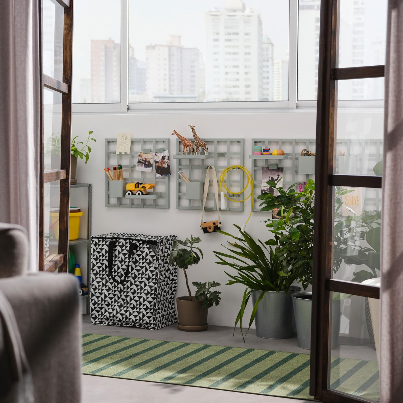 Open doors onto a balcony with grey-green SNICKRA storage boards on a wall, a black/white bag, large potted plants and a rug.