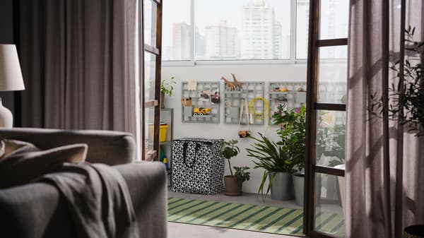 Open doors onto an eclosed balcony with grey-green SNICKRA storage boards on a wall, a black/white bag, large potted plants and a rug.