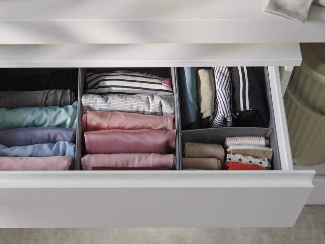 Open drawer with neatly folded clothes organized by color and type in separate compartments, under a white countertop with a basket and folded garment nearby.