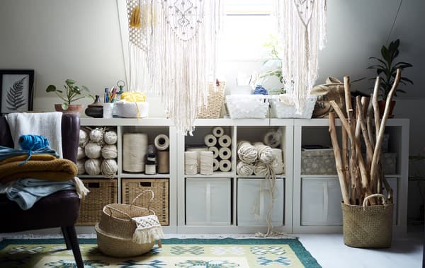 Open storage filled with boxes and creative hobby items, including threads, wooden sticks and hanging macramé items.