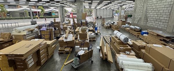 Open warehouse with stacks of cardboard boxes and rolled materials on carts. The industrial setting is busy, with a neutral, organized atmosphere.