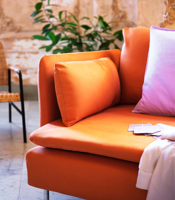 Oranžová pohovka s ružovým vankúšom a svetloružovou prikrývkou. Na pohovke sú položené karty.