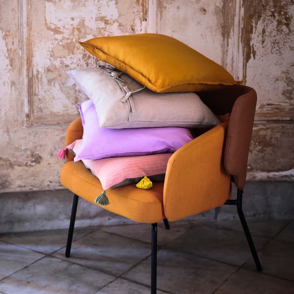 Oranžové kreslo s rozličnými vankúšmi vo fialovej, ružovej a sivej farbe pri béžovej stene vo vidieckom štýle.