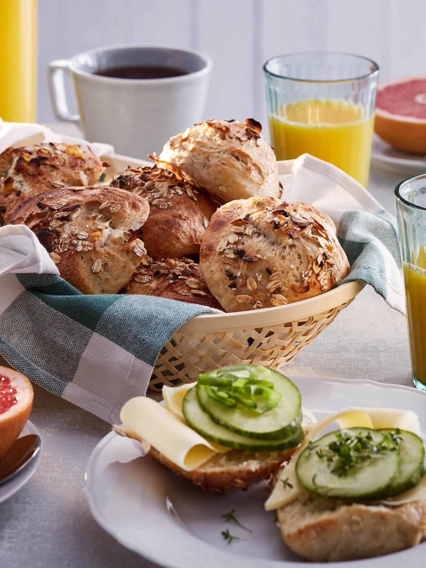 Ošatka vyložená utěrkou plná houstiček, okolo stojí sklenice VARDAGEN s pomerančovým džusem a hrnek s kávou, v popředí je dezertní talíř UPPLAGA s rozkrojenou houstičkou obloženou sýrem a okurkou.