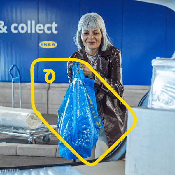 Osoba drži veliku plavu IKEA vrećicu u prostoru za preuzimanje narudžbi s oznakom „Click & collect“. U pozadini su kolica s upakiranim proizvodima i plavi zid s IKEA logotipom.