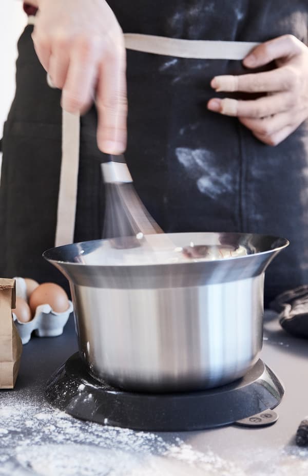 Osoba jednou rukou mieša v miske OMTÄNKSAM s vrchnákom, ktorý sa dá umiestniť pod misku, aby sa nešmýkala.