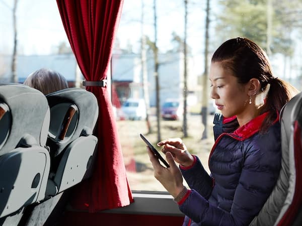 Osoba sjedi u autobusu gledajući u mobitel, pored prozora sa zavjesom.