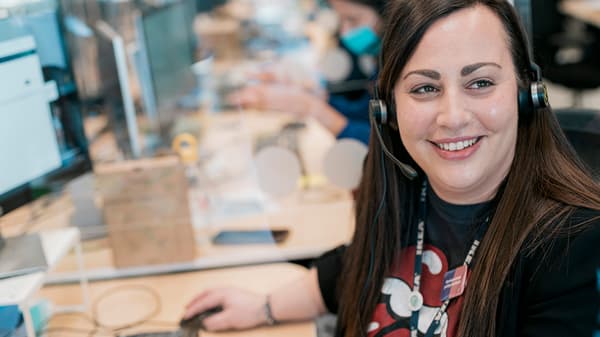 Osoba sjedi za stolom s računalom i slušalicama, u pozadini su drugi ljudi i računala.