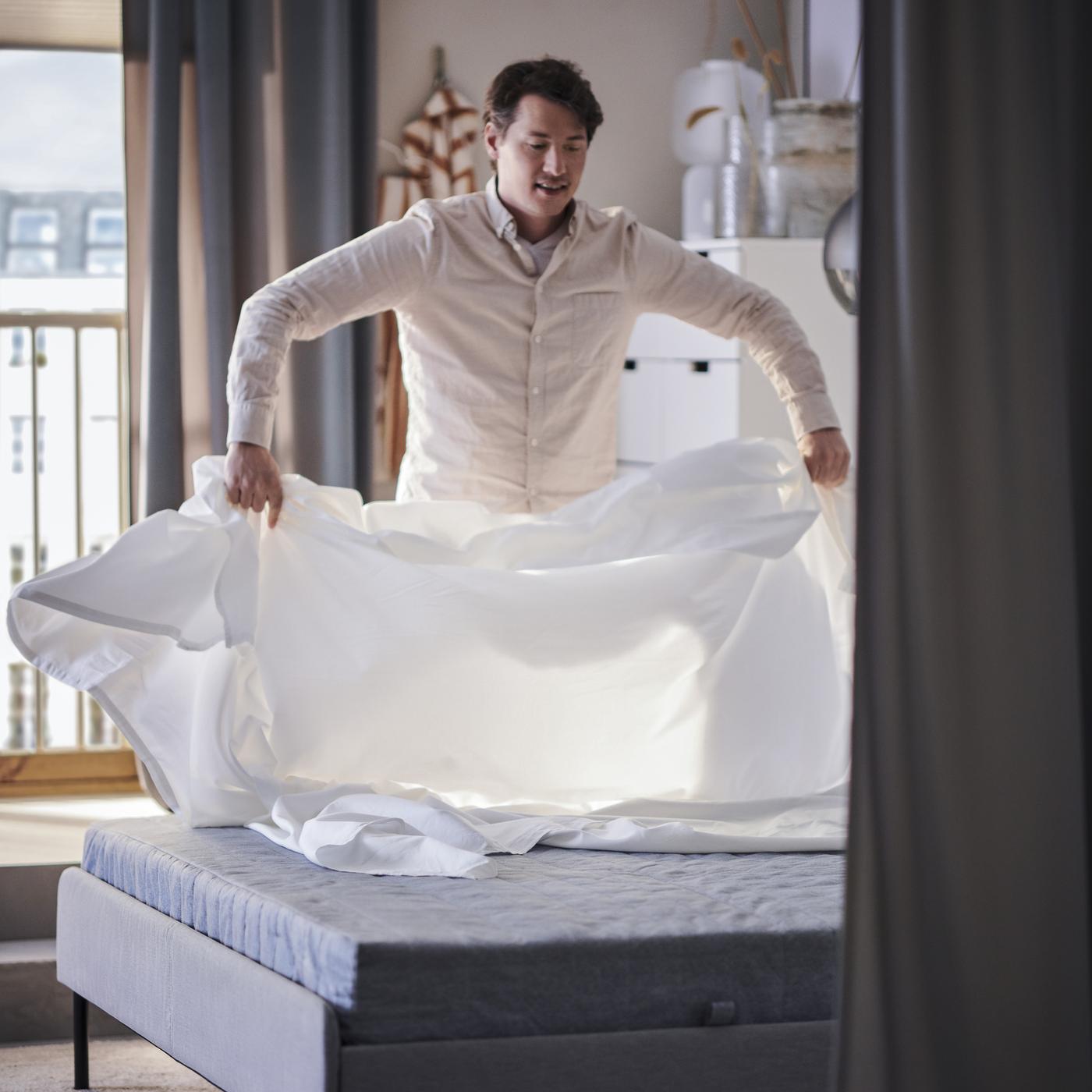 Osoba slaže bijelu plahtu na krevetu u svijetloj spavaćoj sobi.