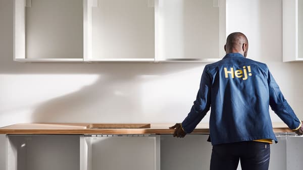 Osoba stoji ispred kuhinjskog elementa u montaži, s drvenom radnom pločom i otvorenim bijelim ormarićima na zidu. Na leđima jakne vidljiv je natpis „Hej!“.