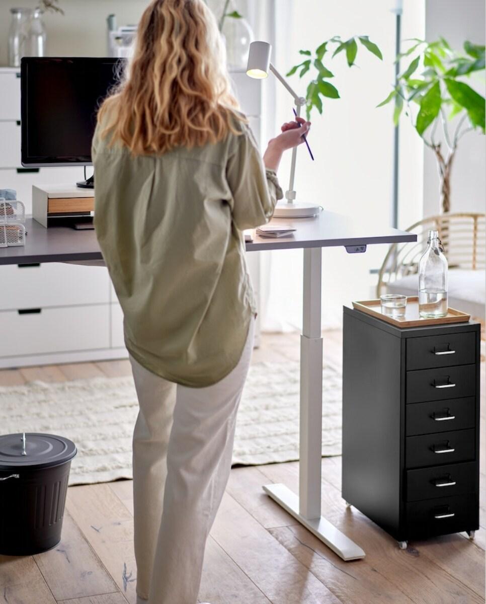 Osoba stoji za radnim stolom, drži lampu, a u pozadini su biljke i uredski namještaj.