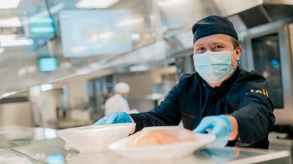 Osoba u zaštitnoj opremi radi u kuhinji i drži pladnjeve s hranom.