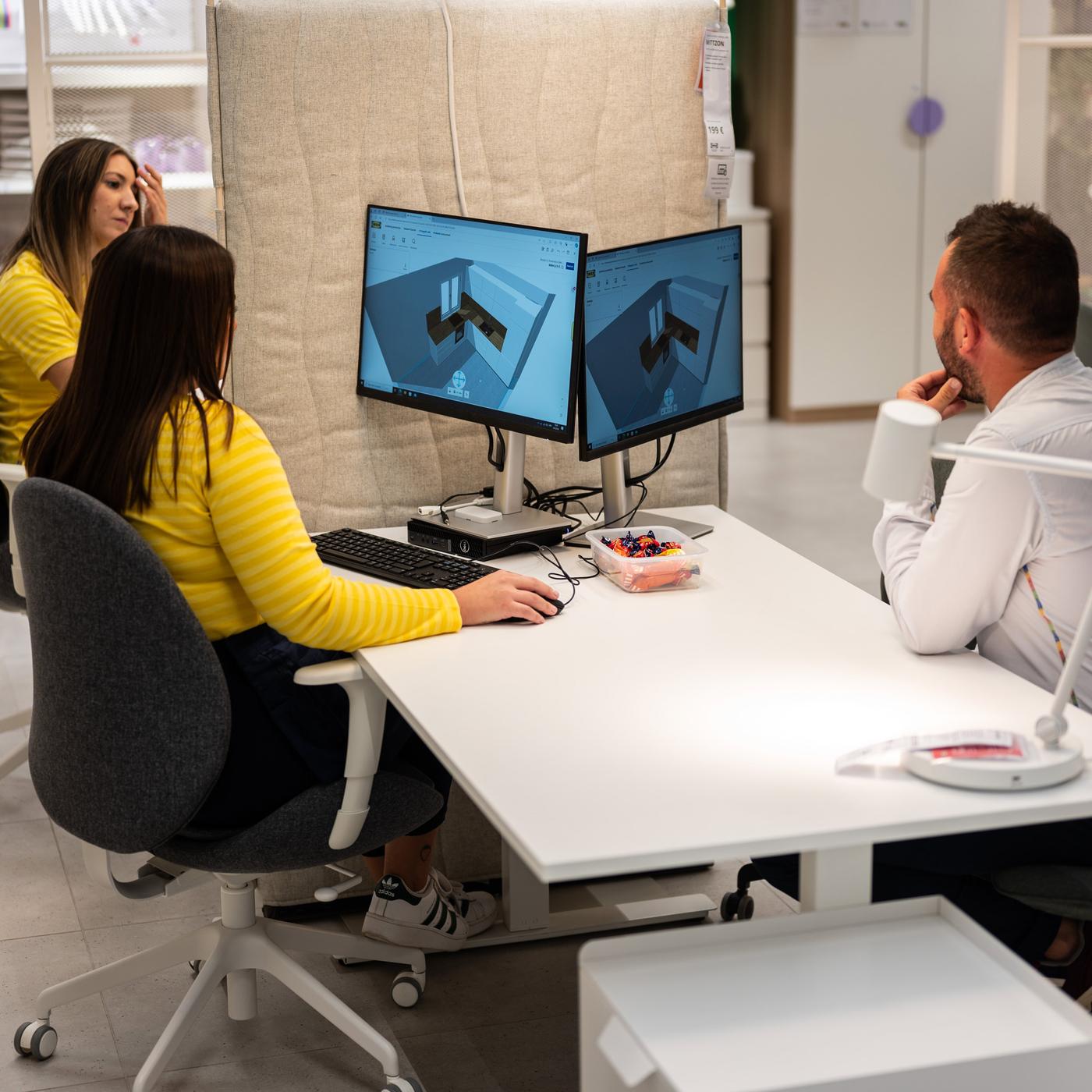 Osoba u žutoj majici sjedi za stolom i radi na računalu s dva monitora, dok dvije osobe sjede nasuprot nje.