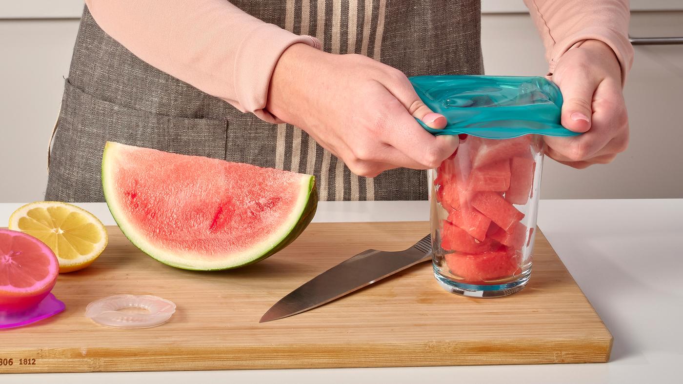 Osoba umiestňuje tyrkysový silikónový kryt na jedlo ÖVERMÄTT na pohár s pokrájaným červeným melónom, ktorý stojí na doske na krájanie.