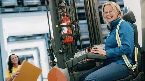 Osoba upravlja viličarem u skladištu, dok druga osoba u pozadini drži žutu mapu.