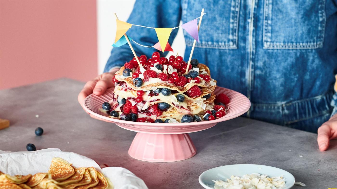 Osoba v džínovém oblečení stojí před pracovní deskou, na které stojí růžový servírovací stojan a dort z palačinek s ovocem. 
