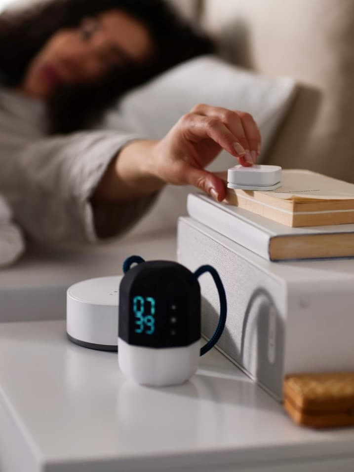 Osoba v posteli siahne po inteligentnom diaľkovom ovládači IKEA STYRBAR na nočnom stolíku vedľa digitálneho budíka IKEA BÅRDRING a reproduktora IKEA SYMFONISK. Moderné doplnky pre inteligentnú domácnosť v spálni.