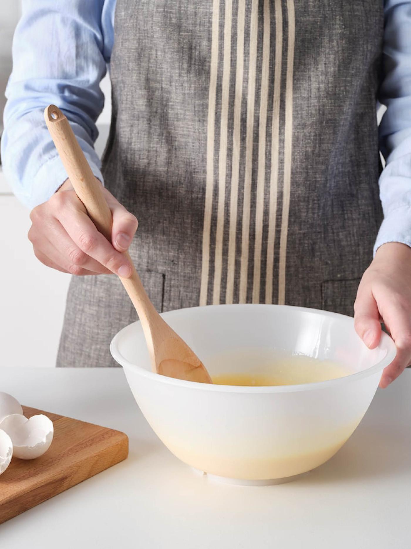 Osoba v zástěře míchá těsto v misce FIKADAGS dřevěnou lžící RÖRT.