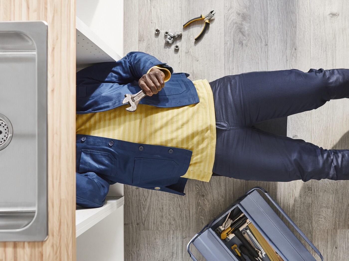 Osoba w niebieskiej kurtce montuje białe meble kuchenne z szafkami stojącymi i wiszącymi oraz laminowanym blatem z imitacją dębu.