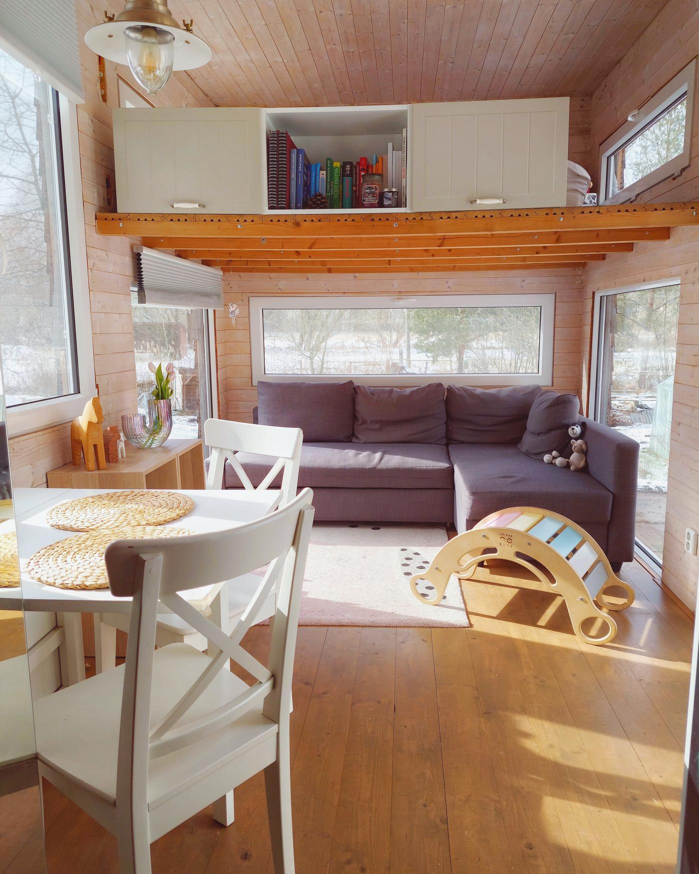 Otevřený obývací pokoj s jídelnou v maringotce, se šedou pohovkou ve tvaru L v pozadí, a bílým jídelním stolem s bílými židlemi INGATORP vepředu. Nad pohovkou je druhé patro sloužící i jako úložné prostory.