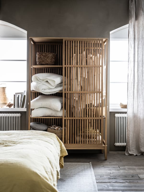 Otvorená skriňa NORDKISA s paplónmi, vankúšmi, dekoráciami a vešiakom na oblečenie medzi dvoma oknami. 