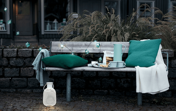 Outdoor, early-evening city setting with a public bench set with a coffee tray, cushions, a throw and decorative lighting.