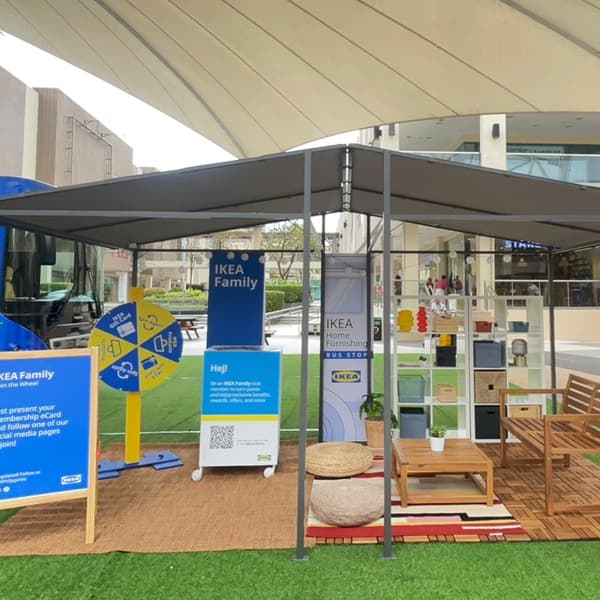 Outdoor IKEA Family booth with blue and yellow signs, a prize wheel, and home decor items under a canopy. Modern, welcoming atmosphere.