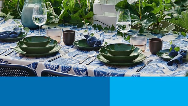 Outdoor table with green dishes, glassware, and blue textiles surrounded by leafy plants.