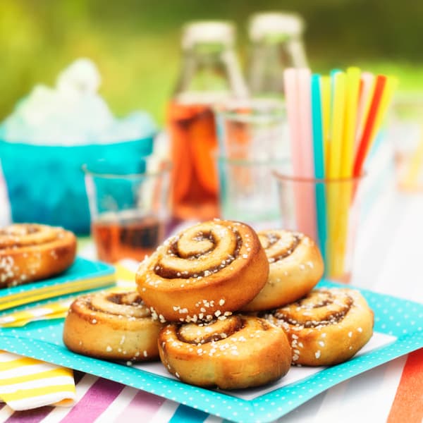 Outdoors, on a table set with bright, summery decorations, there are several cinnamon rolls, colorful straws in a glass, and drinks.