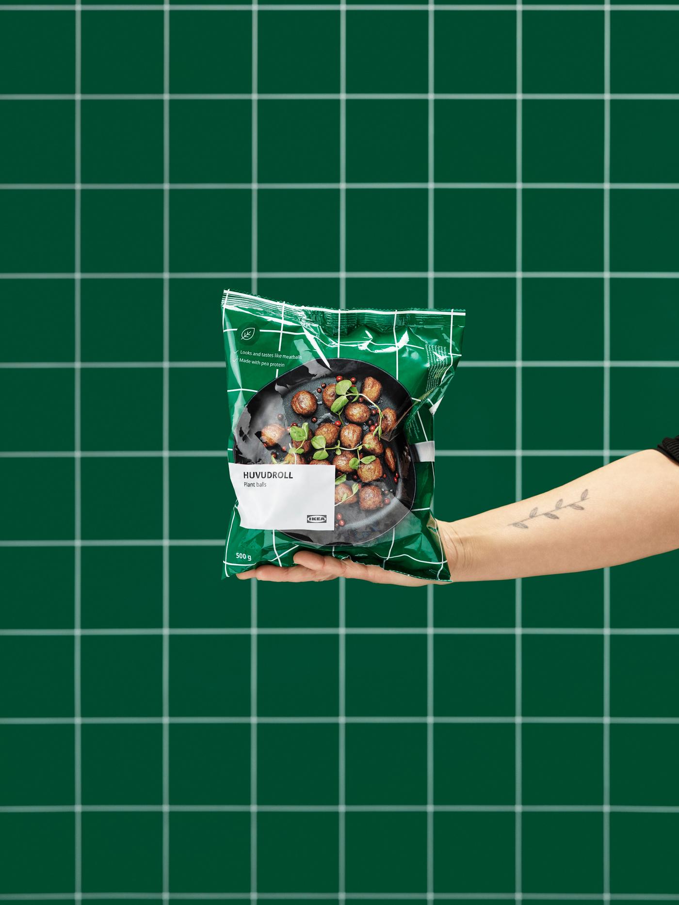 Outstretched hand holding an unopened package of HUVUDROLL veggie balls. Green, tiled wall in the background.