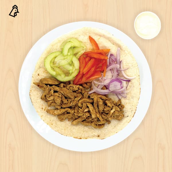 Overhead view of a chicken shawarma wrap on a white plate, topped with cucumber, tomato, red onion, and sauce on a warm wood table.