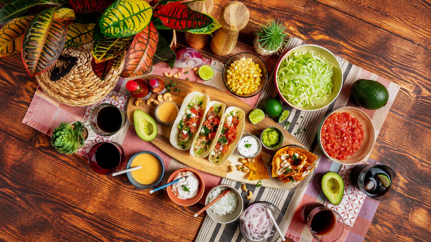 Overhead view of a colourful taco feast on a wooden table featuring soft tacos, tortilla chips with nacho cheese, shredded lettuce, diced tomatoes, avocado, corn, onions, guacamole, sour cream, sauces and refreshing drinks, surrounded by vibrant houseplants.