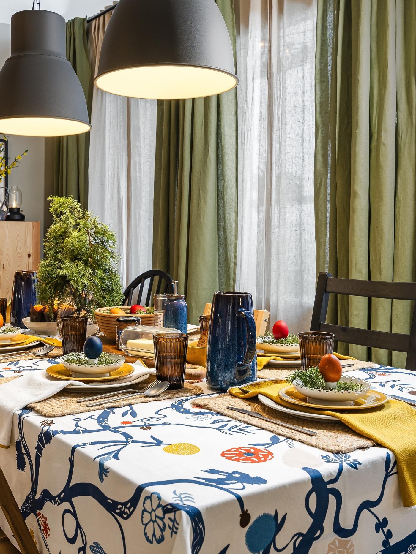 Overhead view of a patterned tablecloth with plates, colourful eggs and simple dinnerware arranged neatly across the table.