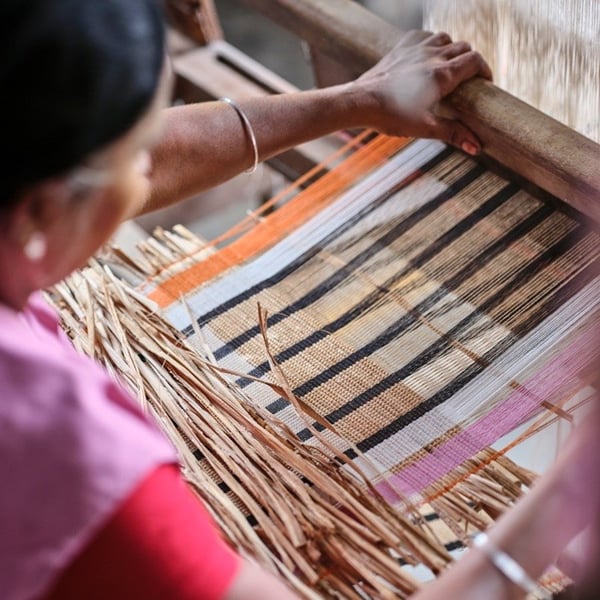 Overhead view of a person weaving a MÄVINN placemat on a handloom, with hands guiding threads to form striped patterns.