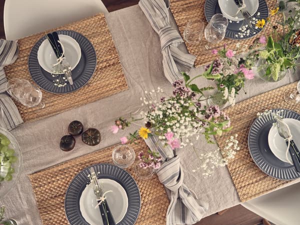 Overhead view of a set dining table with grey plates, white bowls, flowers, glasses and woven placemats.
