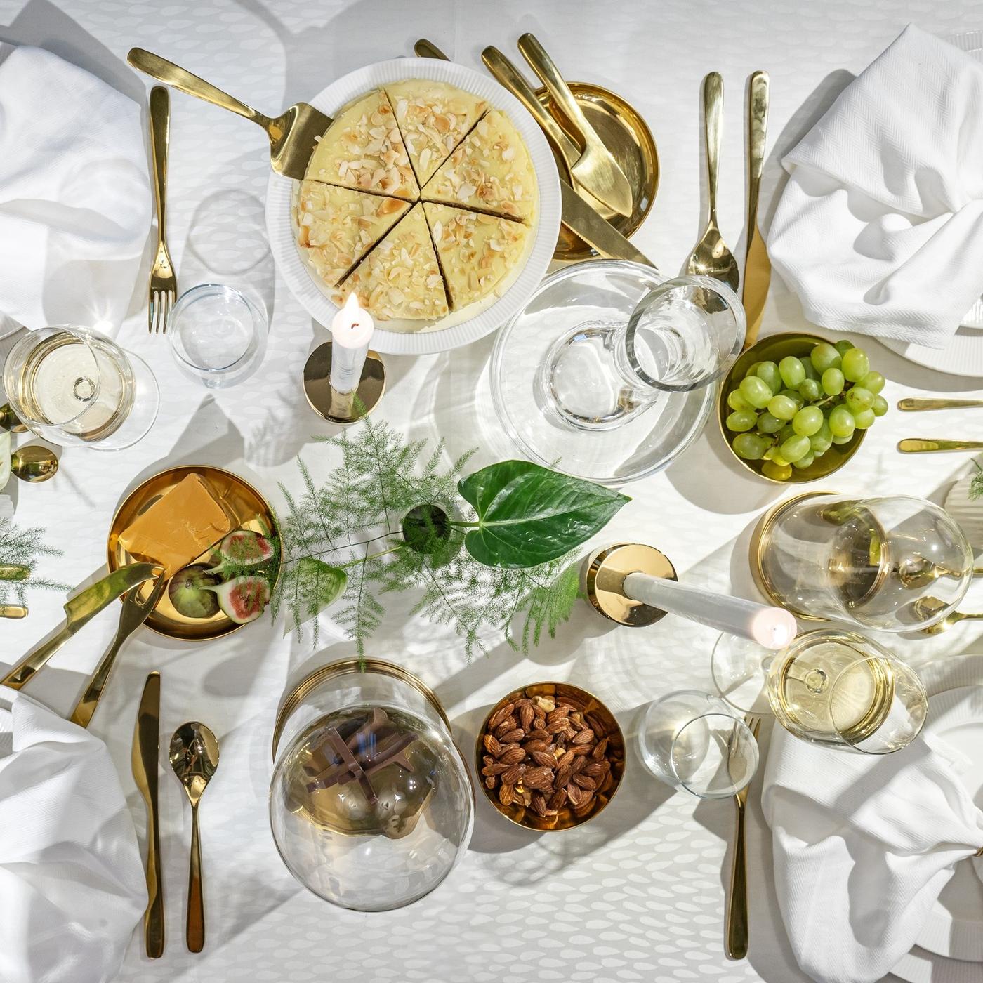 Overhead view of a table setting with layered plates, gold-toned cutlery, clear glasses and small bowls of snacks arranged on a light tablecloth.