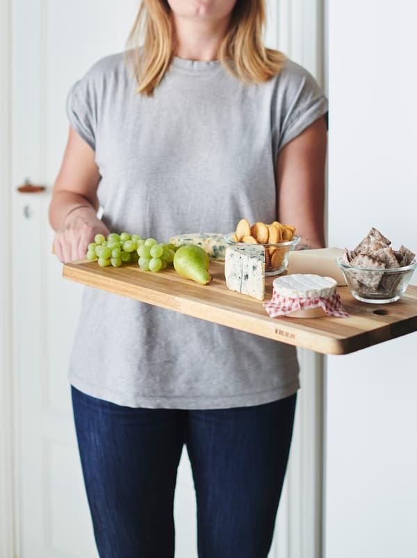 Overkrop og ben af en kvinde i jeans og T-shirt, der rækker et SMÅÄTA skærebræt med oste frem.