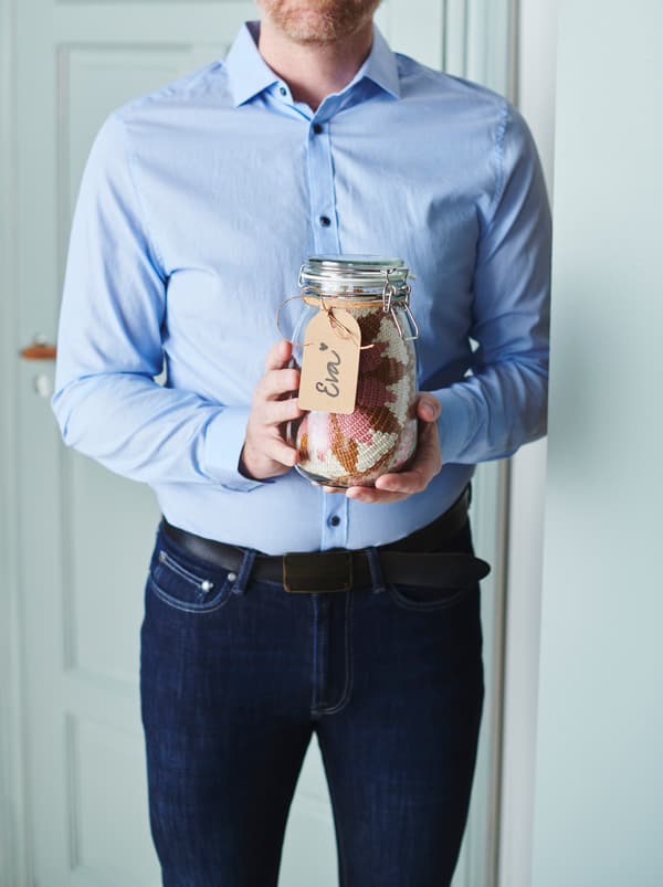 Overkroppen af en mand i jeans og skjorte, der holder en KORKEN krukke af glas med et håndskrevet mærkat og en klud indeni. 