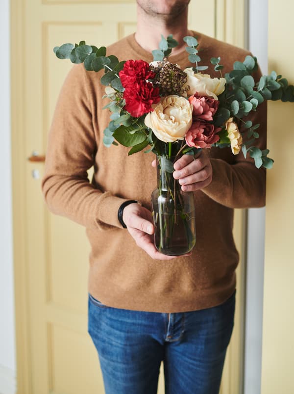Overkroppen af en mand i jeans og sweater, der holder en FÖRENLIG vase med en buket sammensat af f.eks. SMYCKA kunstige blomster. 