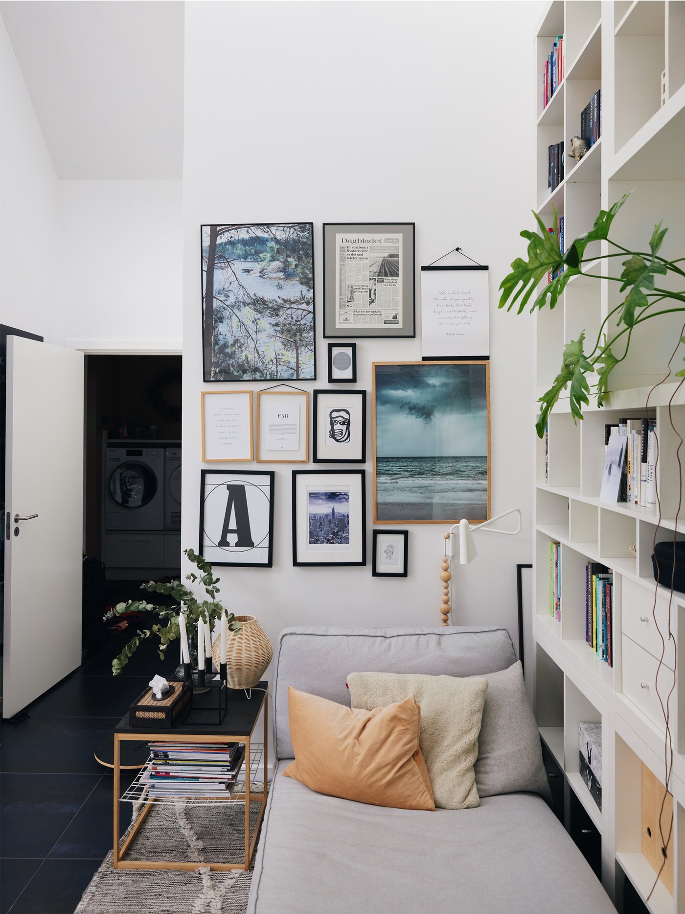 På en gallerivæg hænger en samling af billeder til højre er en reol der fylder hele væggen fyldt med bøger og til venstre står en dør åben.