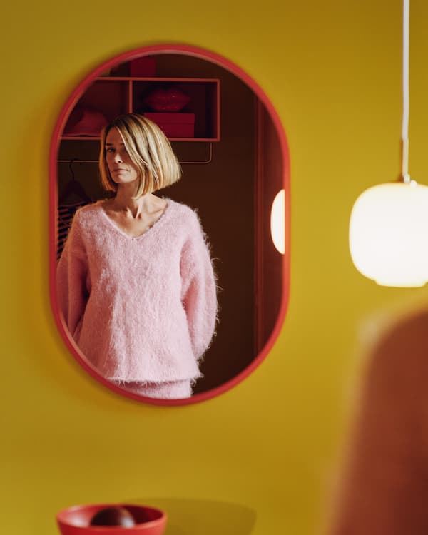 På en gul vägg hänger en spegel med en röd ram. I spegelbilden syns en kvinna i en stickad rosa tröja. 