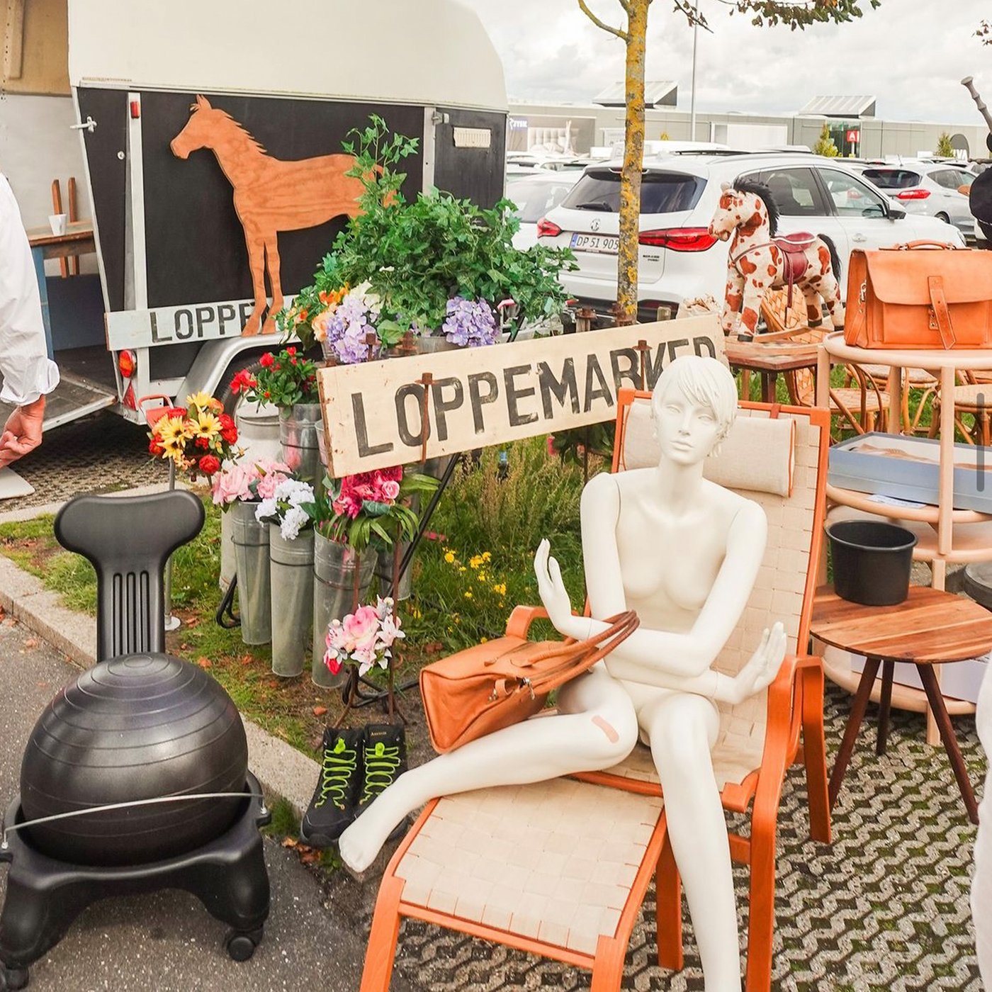 På en liggestol sidder en hvid figur af en kvinde bag figuren hænger et skilt hvor der står loppemarked og rundt omkring skiltet er der fyldt op med forskellige møbler.