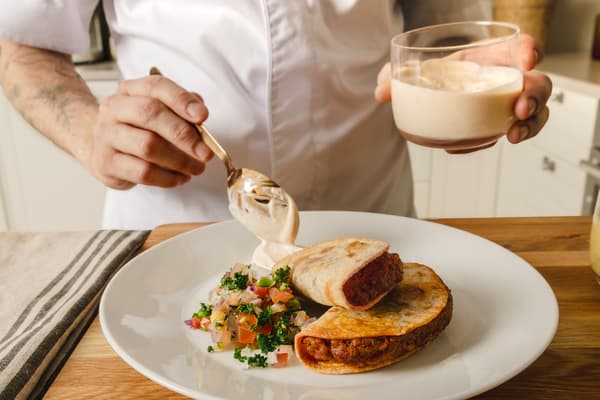 På en vit tallrik ligger två tacos med tillbehör. En person i kockkläder lägger en sked med chilimajonnäs på tallriken.