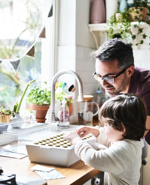 Padre e figlio in una cucina bianca con un kit di idrocoltura VÄXER posto su un bancone in legno di una cucina ben illuminata.