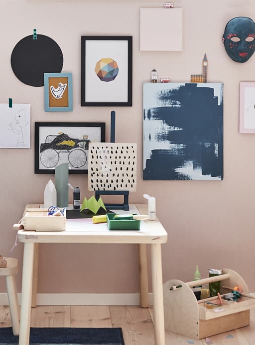 Paintings and drawings displayed on a pink wall above a light wooden desk.