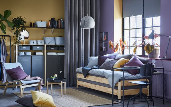 Pangsapuri studio berwarna kuning dan ungu dilengkapi katil sofa UTÅKER berkayu pain yang praktikal yang boleh digunakan sebagai sofa, katil bujang, katil kembar atau katil kelamin. Storan berkayu IVAR dengan unit kabinet berkeluli kelabu boleh dilihat di latar.