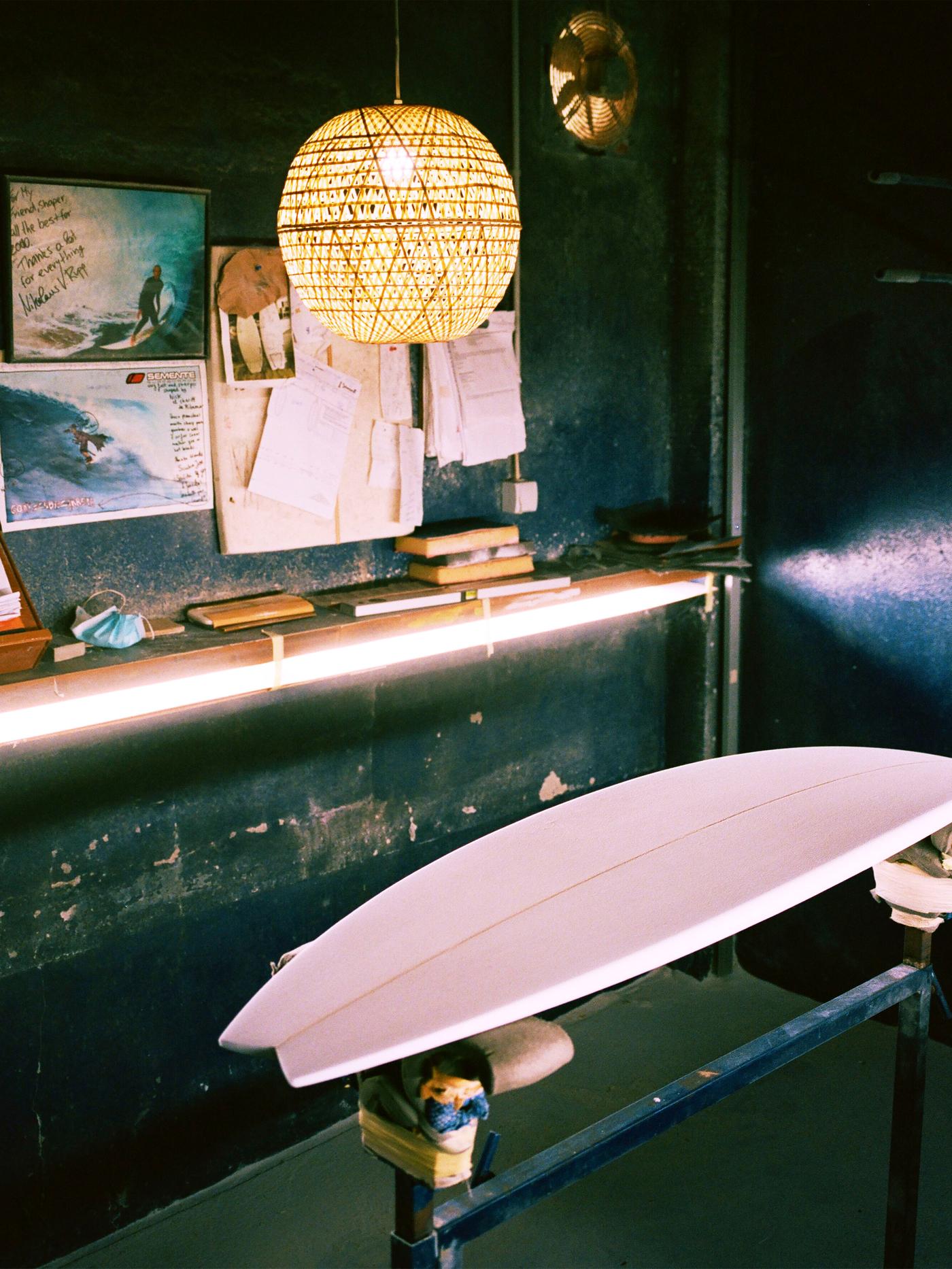 Pantalla para lámpada KÅSEBERGA feita de bambú entretecido colgada sobre unha táboa de surf de madeira situada no taller de cor escura onde traballa o artista.