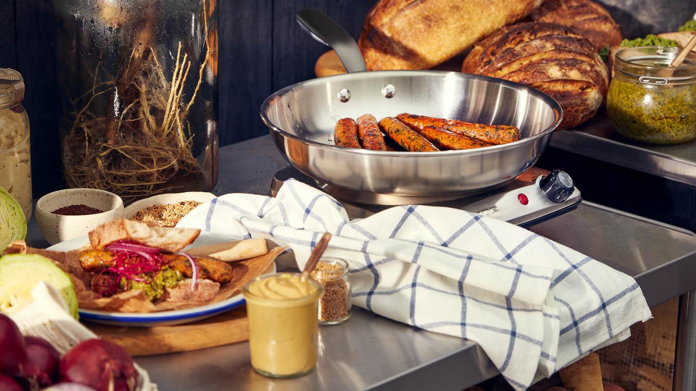 Panvica SENSUELL so zeleninovými hotdogmi KORVMOJ stojí na elektrickom variči, vedľa je položená utierka, tanierik s obloženým hotdogom, pohár s horčicou, červená cibuľa a rozpolená hlávka kapusty, v pozadí je pečivo.