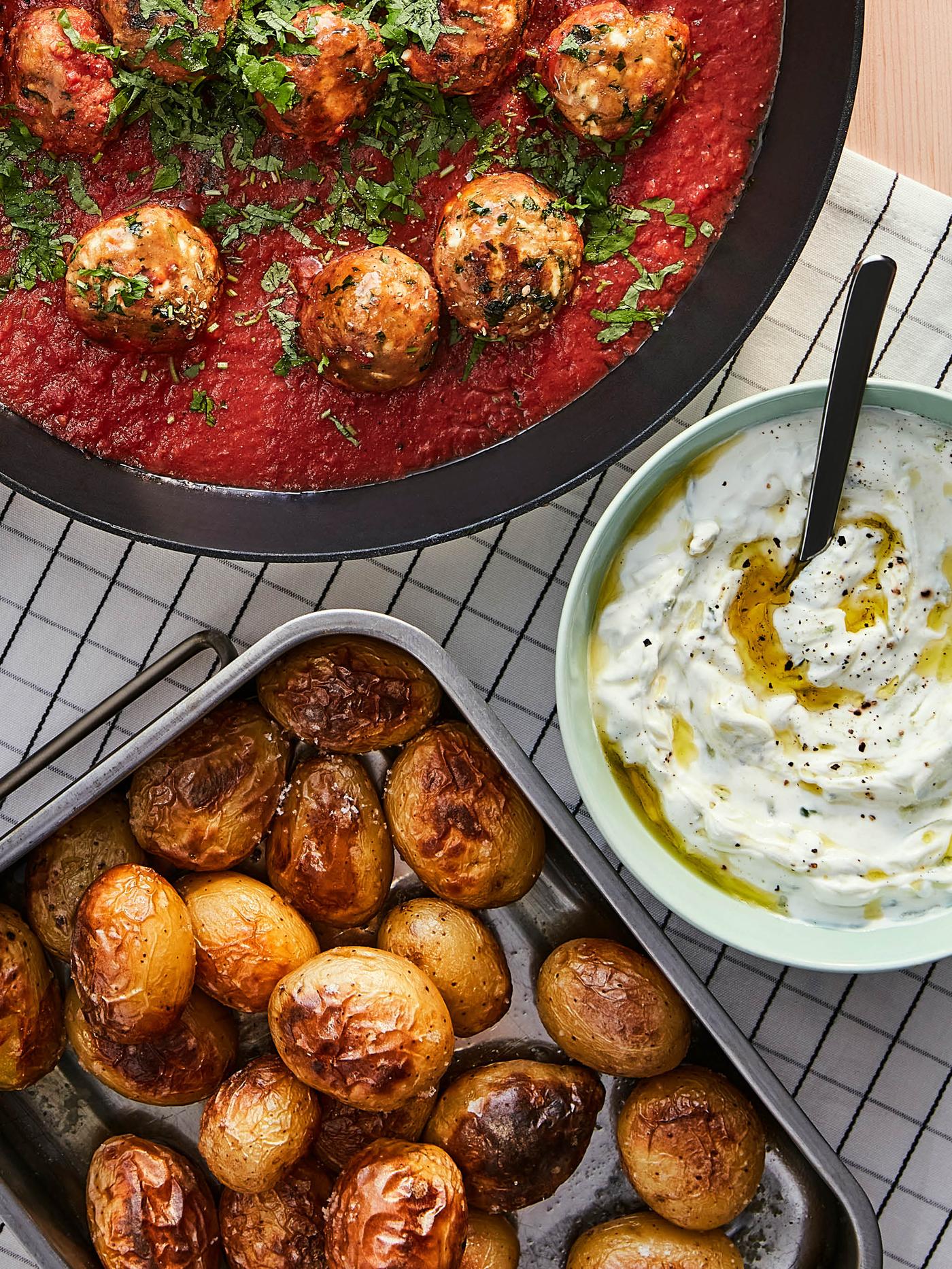 Panvica VARDAGEN s guľôčkami z bezmäsitého mletého VÄRLDSKLOK v paradajkovej omáčke je na stole vedľa svetlozelenej misky s tzatziky a pekáča KONCIS s pečenými zemiakmi.