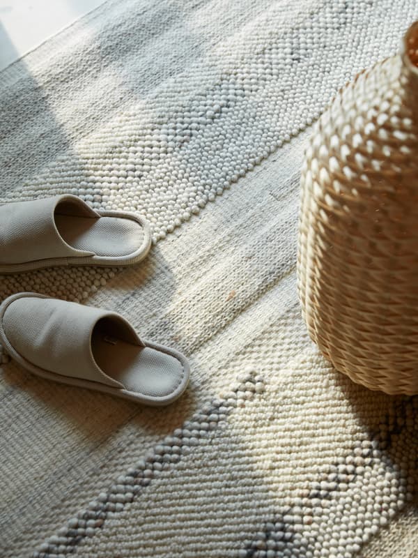 Papuci de casă de culoare crem și o vază din împletitură de bambus pe un covor BRÖNDEN țesut manual.